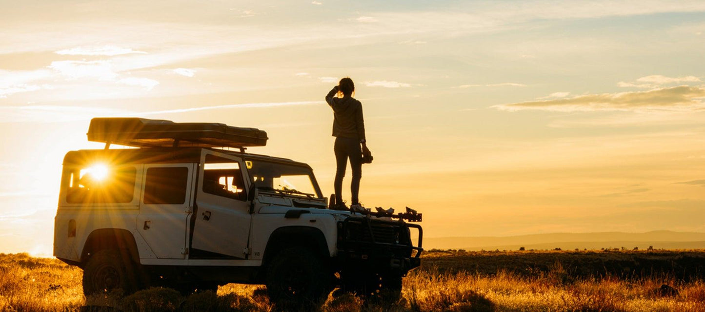 A person stands on top of a white vehicle, silhouetted against a sunset, looking into the distance with a camera.