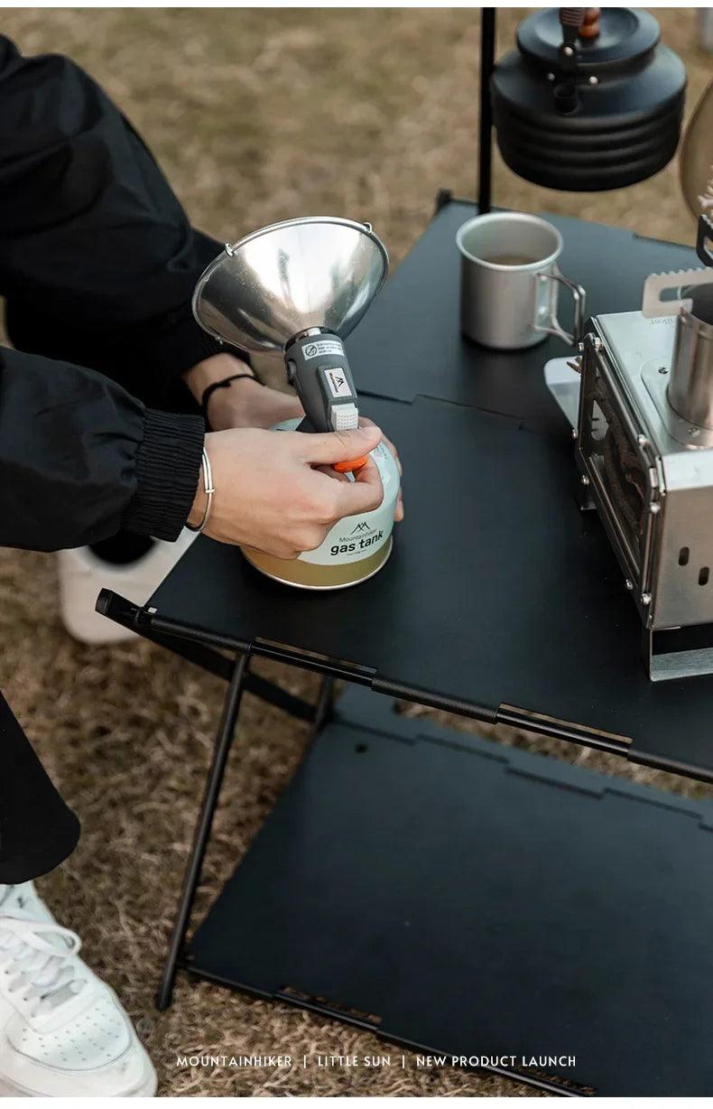 Person holding portable gas lantern on black outdoor table with metal cup and stove, camping scene