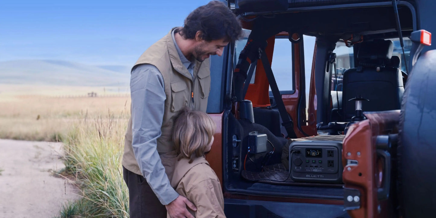 A man and a child are standing next to the BLUETTI Elite 30 V2 Portable Power Station inside a vehicle, smiling.