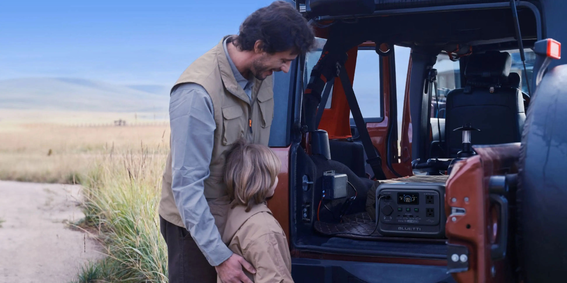 A man and a child are standing next to the BLUETTI Elite 30 V2 Portable Power Station inside a vehicle, smiling.