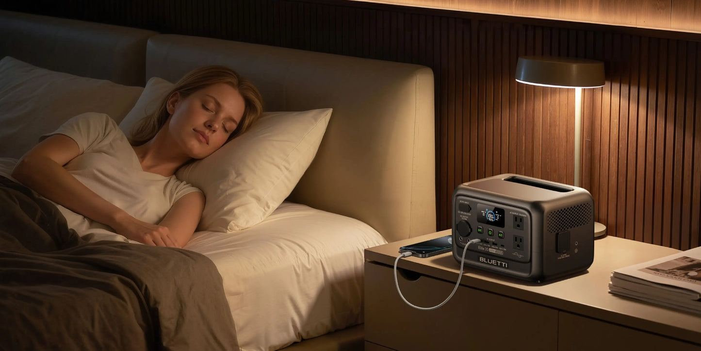 The BLUETTI Elite 30 V2 Portable Power Station features a sleek, compact design, resting on a bedside table. A smartphone is plugged into it while a woman sleeps peacefully nearby.
