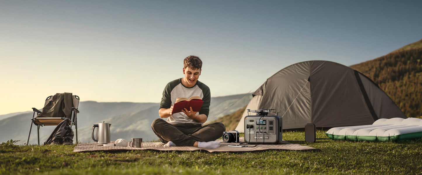 A man sits cross-legged on a picnic blanket, reading a book near the BLUETTI AC50B Portable Power Station, a silver kettle beside him. A camping chair is nearby, with a tent and inflatable mattress in the background.